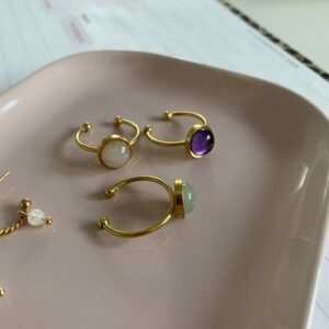 Goudkleurige verstelbare stainlesless steel ring ingelegd met amethist omringd door een ring met rozenkwarts en een ring met aventurijn. De ringen liggen op een roze schaaltje wat op een site tafel staat.