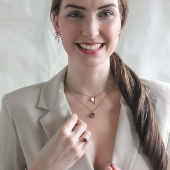 Deze afbeelding toont een vrouw die een goudkleurige stainless steel ketting met amethist steen draagt. De amethist steen in het midden van de ketting voegt een kleurrijk en opvallend element toe aan de outfit van de vrouw. De ketting heeft een fijne schakel en is afgewerkt met een karabijnsluiting. De vrouw draagt de ketting om haar nek en heeft een glimlach op haar gezicht.