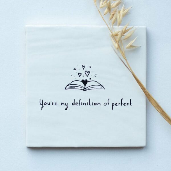 Witte spreukentegel met de tekst "You're my definition of perfect" de tekst is in het zwart geschreven en de afbeelding is een opengeslagen boek waar hartjes uitkomen, dit is in het zwart erop geschilderd.