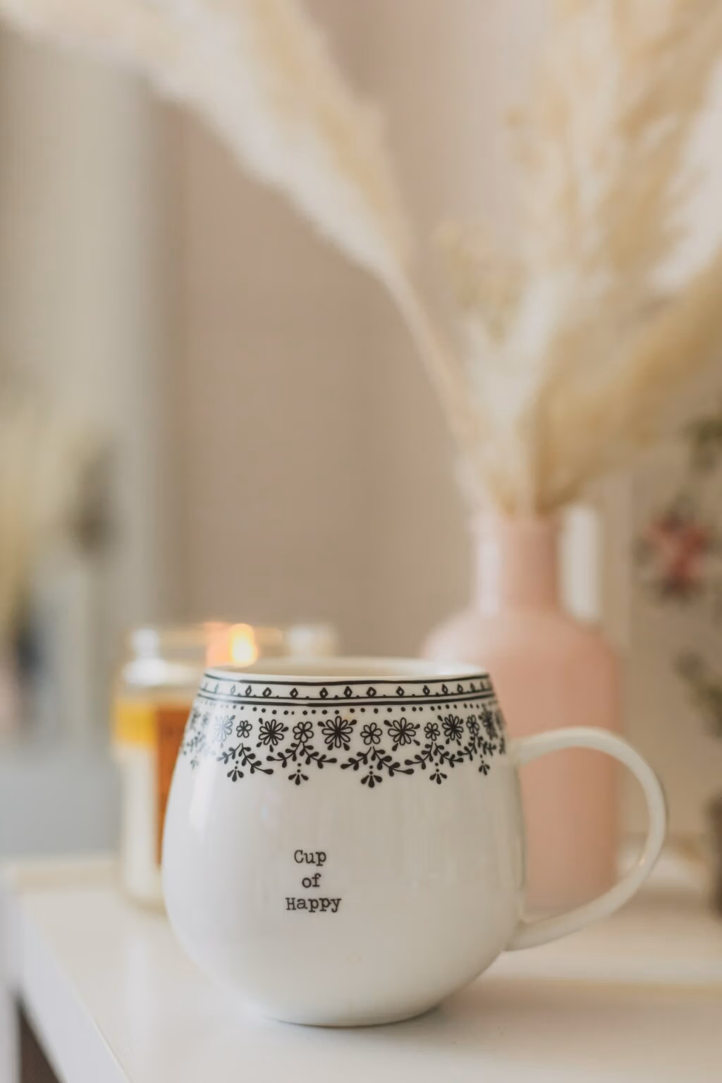 Een witte keramische beker gevuld met warme thee, geserveerd op een houten witte kastje. Den opdruk van de beker is Cup of Happy. Er brand een geurkaars van de Tribeshop.nl, die staat achter de beker.