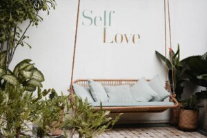 Een rustieke schommelbank omringd door weelderig groen en planten in verschillende tinten groen en bruin. De achtergrond toont een muur met de inspirerende woorden "self love" in sierlijke letters geschreven. De schommelbank biedt een uitnodigende plek om te ontspannen en te genieten van de natuurlijke omgeving terwijl de boodschap op de muur eraan herinnert om tijd te nemen voor zelfliefde en zorg.