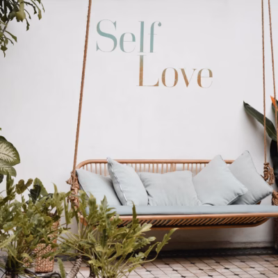Een rustieke schommelbank omringd door weelderig groen en planten in verschillende tinten groen en bruin. De achtergrond toont een muur met de inspirerende woorden "self love" in sierlijke letters geschreven. De schommelbank biedt een uitnodigende plek om te ontspannen en te genieten van de natuurlijke omgeving terwijl de boodschap op de muur eraan herinnert om tijd te nemen voor zelfliefde en zorg.
