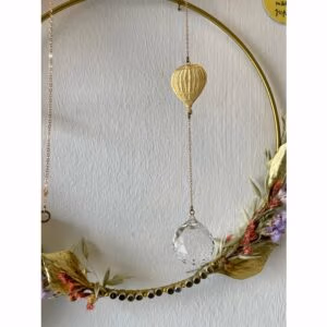 Suncatcher met een decoratieve goudkleurige ballon op de achtergrond een crèmekleurige muur met een decoratieve krans.