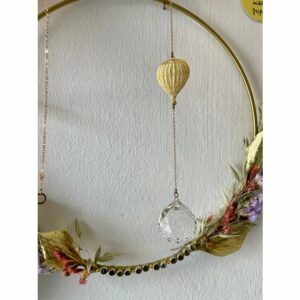 Suncatcher met een decoratieve goudkleurige ballon op de achtergrond een crèmekleurige muur met een decoratieve krans.
