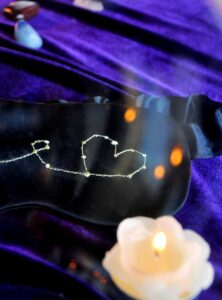 Zwart Satijnen slaapmasker met handen en een hart erop geborduurd. Het slaapmasker ligt op een velvet blauwpaarse achtergrond en er staat een witte kaars naast die brand.