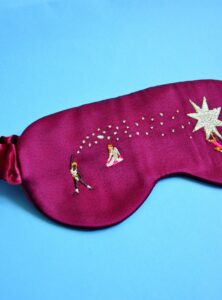 Bordeaux rode satijnen slaapmasker met 3 vrouwelijke illustraties en sterren erop geborduurd op. Het slaapmasker rust op een turquoise ondergrond.