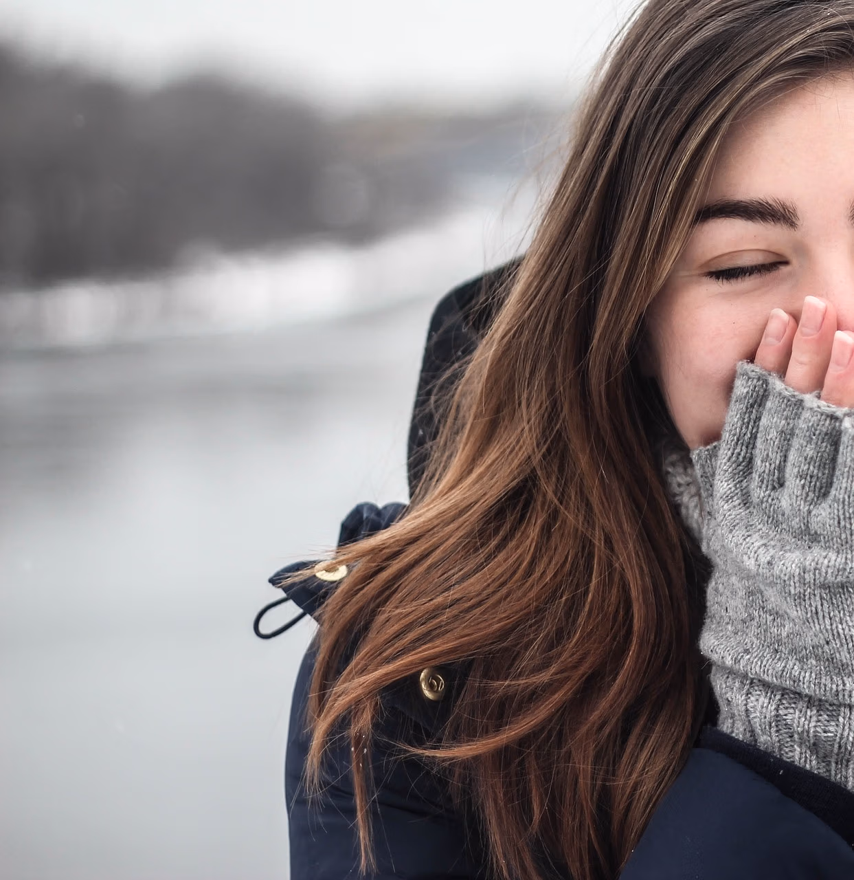 Vrouw met bruin haar in een winterjas en handschoenen staat in een serene sneeuwlandschap met haar ogen gesloten, genietend van de rust.