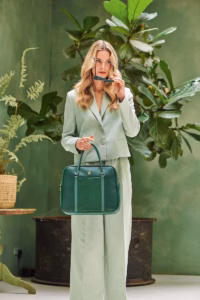 Vrouw met lang, golvend blond haar in een chique lichtgroen pak, staand in een groene ruimte met planten, houdt de Pip Studio Schoudertas Groen elegant in haar hand.