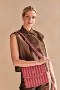 Blonde vrouw in een bruine outfit draagt de Pip Studio Crossbody-Tas Roze/Bordeaux met gestreept design, goudkleurige details en een verstelbare schouderband met geborduurde bloemenprint.