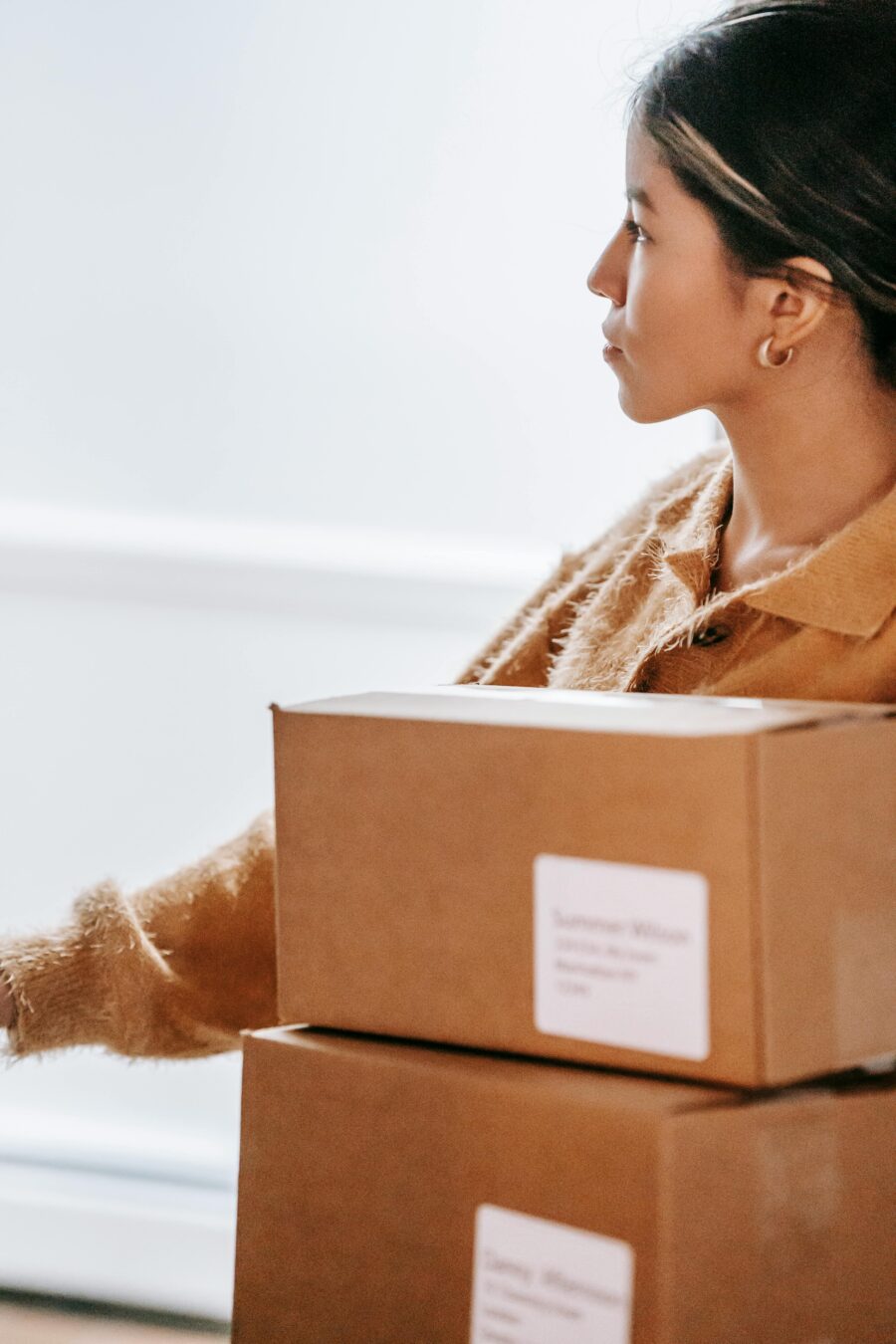 Crop concentrated Asian female in warm sweater standing with carton boxes in light post office and looking away thoughtfully